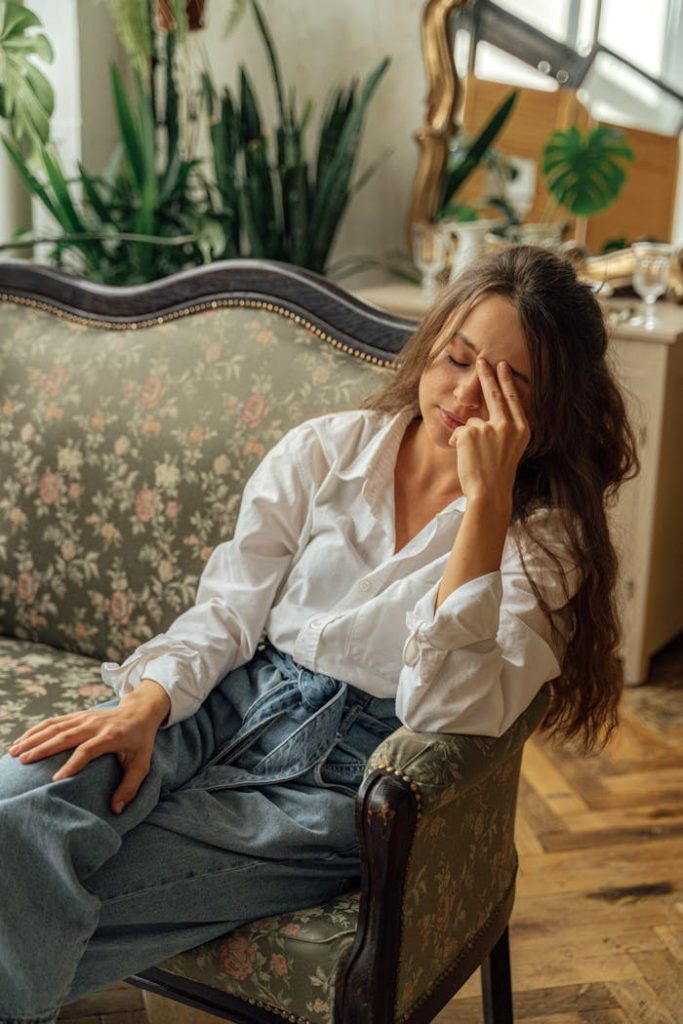 woman-in-white-dress-shirt-and-blue-denim-jeans-sitting-on-couch-5217843 A woman relaxes on a floral vintage sofa in a cozy, light-filled room with plants.